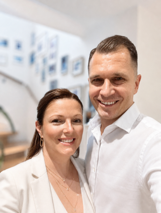 Michael & Sabrina Männing stehen in einem hellen Geschäftsraum nebeneinander und lächeln. (Vollbild)